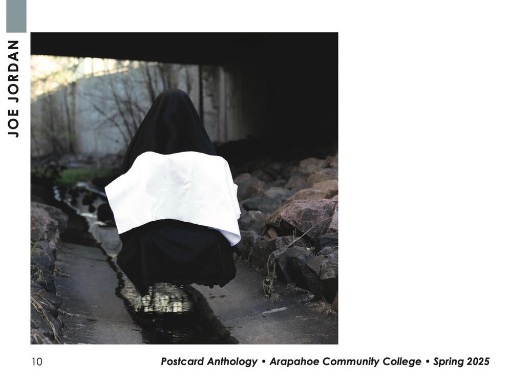 The photograph shows a figure covered head to toe in a black covering with a white blanket resting on its lab. The figure is sitting under a dark bridge.